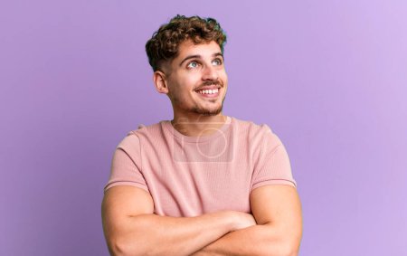 Photo for Young adult caucasian man feeling happy, proud and hopeful, wondering or thinking, looking up to copy space with crossed arms - Royalty Free Image
