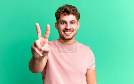 Foto de Young adult caucasian man smiling and looking happy, carefree and positive, gesturing victory or peace with one hand - Imagen libre de derechos