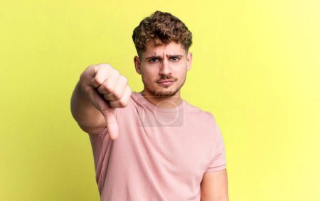 Foto de Young adult caucasian man feeling cross, angry, annoyed, disappointed or displeased, showing thumbs down with a serious look - Imagen libre de derechos