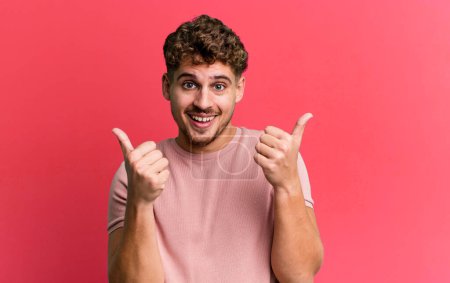 Téléchargez les photos : Young adult caucasian man smiling joyfully and looking happy, feeling carefree and positive with both thumbs up - en image libre de droit