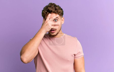 Photo for Young adult caucasian man looking shocked, scared or terrified, covering face with hand and peeking between fingers - Royalty Free Image