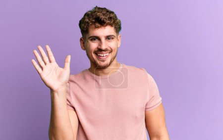 Téléchargez les photos : Young adult caucasian man smiling happily and cheerfully, waving hand, welcoming and greeting you, or saying goodbye - en image libre de droit