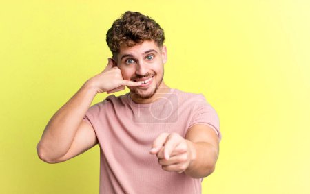 Téléchargez les photos : Young adult caucasian man smiling cheerfully and pointing to camera while making a call you later gesture, talking on phone - en image libre de droit
