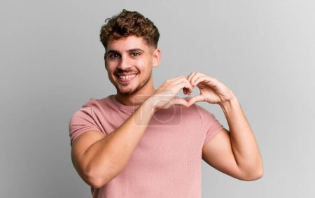 Foto de Young adult caucasian man smiling and feeling happy, cute, romantic and in love, making heart shape with both hands - Imagen libre de derechos