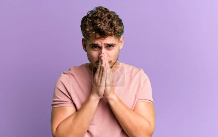 young adult caucasian man feeling worried, hopeful and religious, praying faithfully with palms pressed, begging forgiveness