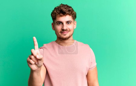 Photo for Young adult caucasian man smiling and looking friendly, showing number one or first with hand forward, counting down - Royalty Free Image