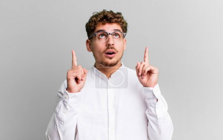 Téléchargez les photos : Young adult caucasian man looking shocked, amazed and open mouthed, pointing upwards with both hands to copy space - en image libre de droit
