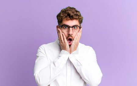 Photo for Young adult caucasian man feeling shocked and scared, looking terrified with open mouth and hands on cheeks - Royalty Free Image