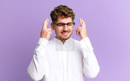Photo for Young adult caucasian man smiling and anxiously crossing both fingers, feeling worried and wishing or hoping for good luck - Royalty Free Image