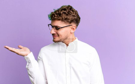 Photo for Young adult caucasian man feeling happy and smiling casually, looking to an object or concept held on the hand on the side - Royalty Free Image