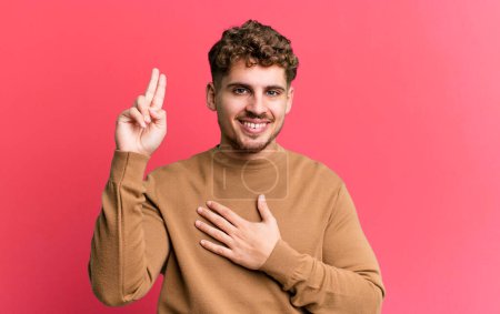 Photo for Young adult caucasian man looking happy, confident and trustworthy, smiling and showing victory sign, with a positive attitude - Royalty Free Image
