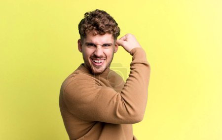 Téléchargez les photos : Young adult caucasian man feeling happy, satisfied and powerful, flexing fit and muscular biceps, looking strong after the gym - en image libre de droit