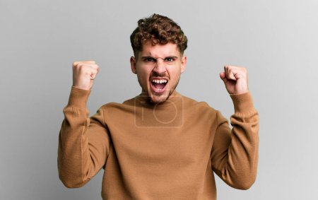 Photo for Young adult caucasian man shouting aggressively with an angry expression or with fists clenched celebrating success - Royalty Free Image