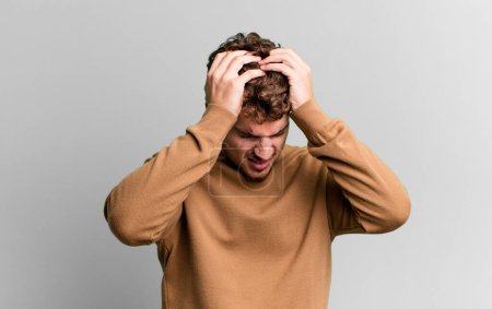 Foto de Young adult caucasian man feeling stressed and frustrated, raising hands to head, feeling tired, unhappy and with migraine - Imagen libre de derechos