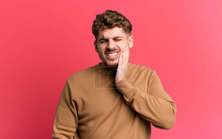 Foto de Young adult caucasian man holding cheek and suffering painful toothache, feeling ill, miserable and unhappy, looking for a dentist - Imagen libre de derechos