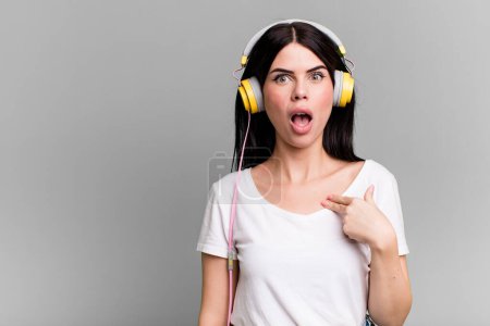 Foto de Mirando sorprendido y sorprendido con la boca abierta, apuntando hacia sí mismo. escuchar música qith a auriculares - Imagen libre de derechos