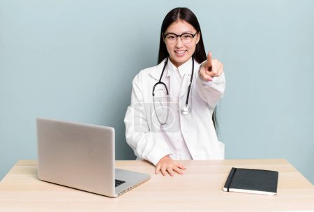 Photo for Pretty asian woman pointing at camera choosing you. physician desk and laptop - Royalty Free Image