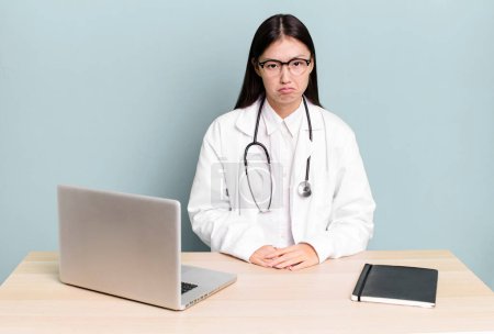 Photo for Pretty asian woman feeling sad and whiney with an unhappy look and crying. physician desk and laptop - Royalty Free Image