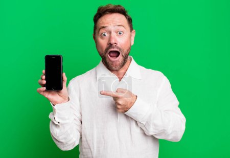 Photo for Middle age man looking excited and surprised pointing to the side. showing a smartphone - Royalty Free Image