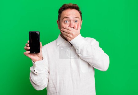 Foto de Hombre de mediana edad cubriendo la boca con las manos con un shock. mostrar un smartphone - Imagen libre de derechos