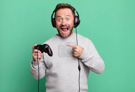 Téléchargez les photos : Homme d'âge moyen regardant excité et surpris pointant vers le côté. concept gamer avec un contrôle et un casque - en image libre de droit