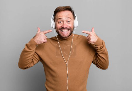 Téléchargez les photos : Homme d'âge moyen souriant avec confiance pointant vers son propre sourire large. écouter de la musique avec un casque - en image libre de droit