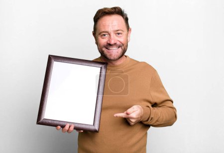 Photo for Middle age man smiling cheerfully, feeling happy and pointing to the side with an empty frame - Royalty Free Image