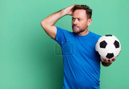 Foto de Hombre de mediana edad sintiéndose desconcertado y confundido, rascándose la cabeza. con una pelota de fútbol. concepto de fitness - Imagen libre de derechos