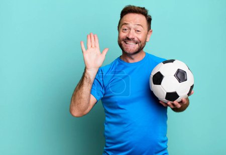 Foto de Hombre de mediana edad sonriendo felizmente, saludándote con la mano, dándote la bienvenida y saludándote. con una pelota de fútbol. concepto de fitness - Imagen libre de derechos