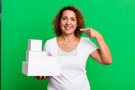 Photo for Pretty middle age woman smiling confidently pointing to own broad smile. blank white boxes packaging - Royalty Free Image