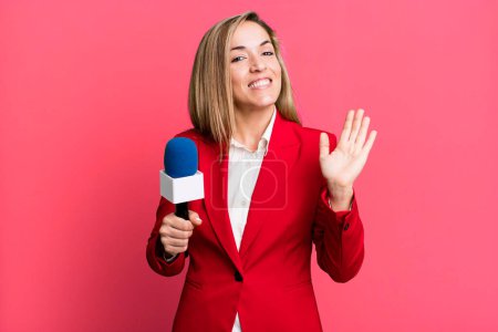 Photo for Pretty blonde woman smiling happily, waving hand, welcoming and greeting you. presenter with a microphone concept - Royalty Free Image
