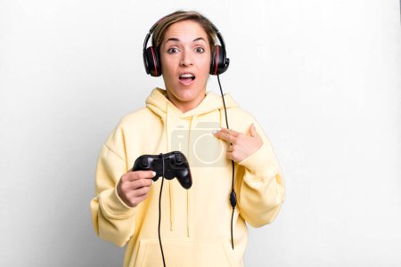 Téléchargez les photos : Pretty blonde woman looking shocked and surprised with mouth wide open, pointing to self. gamer with headset and a controller - en image libre de droit