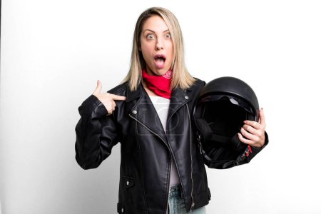 Foto de Pretty blonde woman looking shocked and surprised with mouth wide open, pointing to self. motorbike rider and helmet concept - Imagen libre de derechos