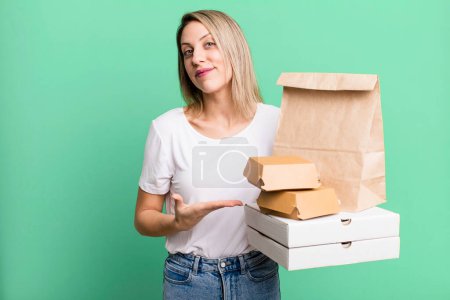 Photo for Pretty blonde woman smiling cheerfully, feeling happy and showing a concept. paper fast food take away packages - Royalty Free Image