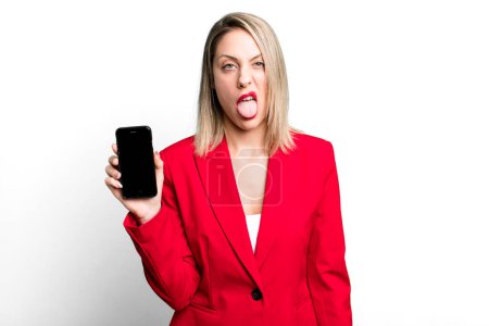 Foto de Bonita mujer rubia sintiéndose disgustada e irritada y la lengua hacia fuera. empresaria mostrando una pantalla de teléfono - Imagen libre de derechos