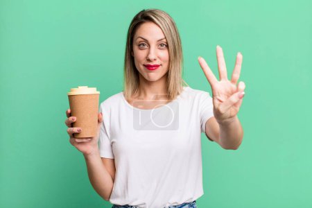 Foto de Bonita mujer rubia sonriendo y buscando amigable, mostrando el número tres con un café caliente - Imagen libre de derechos