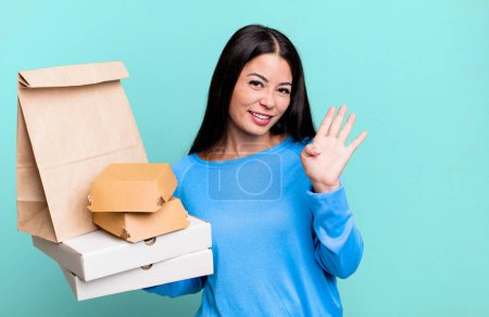 Photo for Hispanic pretty woman smiling happily, waving hand, welcoming and greeting you. with take away fast food packages - Royalty Free Image