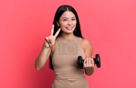 Foto de Hispanic pretty woman smiling and looking friendly, showing number two. fitness concept and dumbbell - Imagen libre de derechos