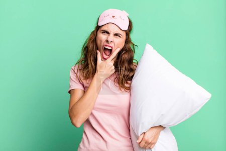 Photo for Hispanic pretty woman with mouth and eyes wide open and hand on chin wearing pajamas and a pillow - Royalty Free Image