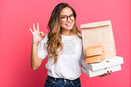 Photo for Hispanic pretty woman feeling happy, showing approval with okay gesture. with fast food packages - Royalty Free Image