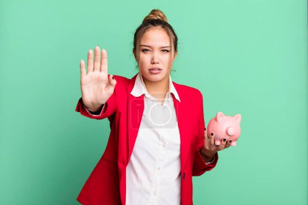 Photo for Hispanic pretty woman looking serious showing open palm making stop gesture with a piggy bank - Royalty Free Image