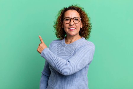 Foto de Mujer hispana de mediana edad sonriendo alegremente, sintiéndose feliz y señalando hacia un lado y hacia arriba, mostrando el objeto en el espacio de copia - Imagen libre de derechos