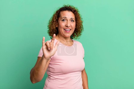 Foto de Mujer hispana de mediana edad sintiéndose feliz, divertida, segura, positiva y rebelde, haciendo letrero de rock o heavy metal con la mano - Imagen libre de derechos