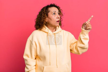 Photo for Middle age hispanic woman feeling shocked and surprised, pointing and looking upwards in awe with amazed, open-mouthed look - Royalty Free Image