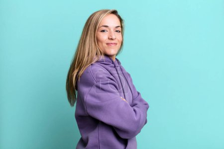 Téléchargez les photos : Blonde adult woman smiling to camera with crossed arms and a happy, confident, satisfied expression, lateral view - en image libre de droit