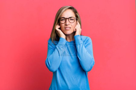 Photo for Blonde adult woman looking angry, stressed and annoyed, covering both ears to a deafening noise, sound or loud music - Royalty Free Image