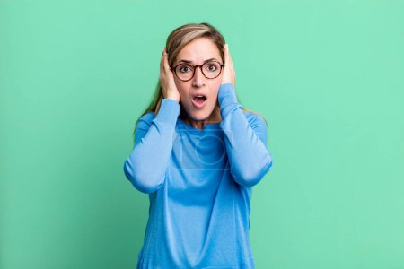 Photo for Blonde adult woman looking unpleasantly shocked, scared or worried, mouth wide open and covering both ears with hands - Royalty Free Image