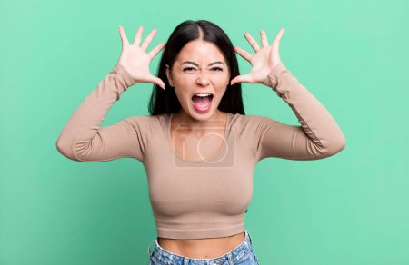 Photo for Pretty latin woman screaming in panic or anger, shocked, terrified or furious, with hands next to head - Royalty Free Image