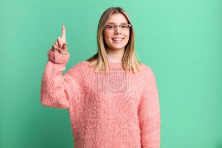 Photo for Blonde pretty woman smiling cheerfully and happily, pointing upwards with one hand to copy space - Royalty Free Image