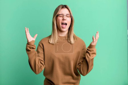 Photo for Blonde pretty woman furiously screaming, feeling stressed and annoyed with hands up in the air saying why me - Royalty Free Image